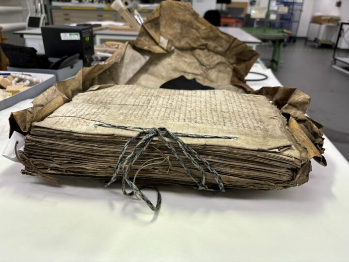 A large bundle of papers stitched together with blue and white cord sits on a table. These papers are surrounded by a crumpled parchment cover.