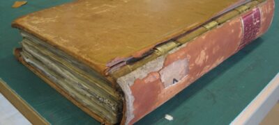 A closed book on a table with damage to its spine, which is detaching from the front cover and has lost areas of its covering material.