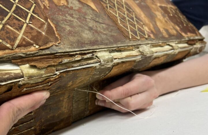Two hands support the spine of a large, open book. One hand is pulling a sewing thread through the spine of the book, to re-attach a loose page within the bookblock. 
