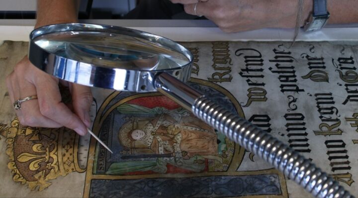 A hand, holding a small wooden tool, hovers over a decoratively painted manuscript, with a magnifying glass in the foreground.