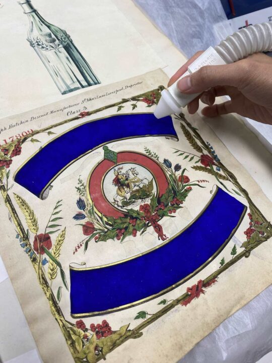 A square handkerchief showing the Emperor Napoleon riding a horse in the centre. Surrounding them are colourful borders, including bright blue scroll borders above and below the image. A conservator's hand holds a white tube over the handkerchief. This has adhesive in it and is being used to stabilise the powdery pigment on the top blue border.
