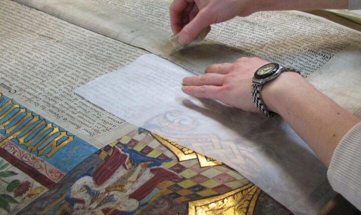 Two hands are on top of an illuminated parchment membrane. The right hand is holding a sponge and cleaning areas of the parchment where there is no text or images. The left hand holds the parchment in place. A transparent piece of tissue lies over the illumination to protect it.