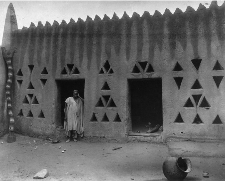 A person stands in the doorway of a stone building. Ornamental windows in the shape of small triangles are cut into the side of the building, creating geometric shapes.