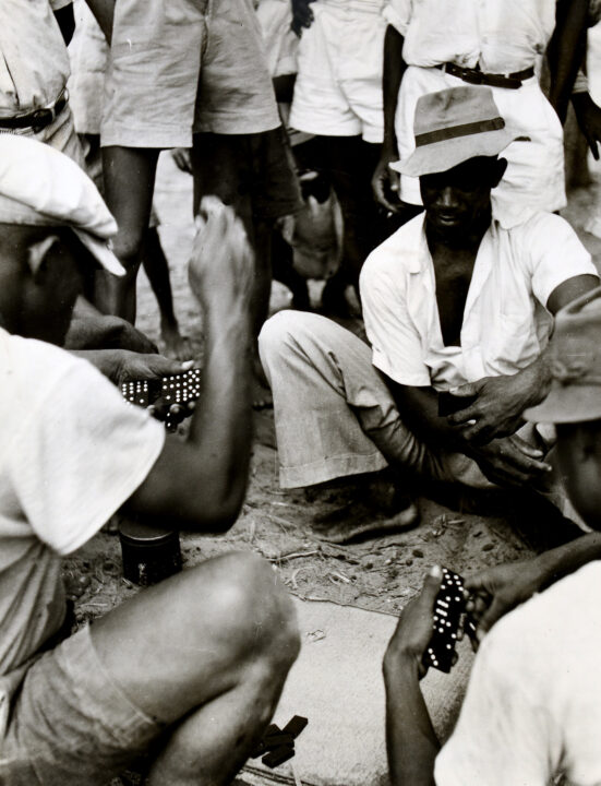 Two St Lucian men crouching on the ground and playing a game of dominoes. They wear white shirts and hats. They are surrounded by a crowd of onlookers.