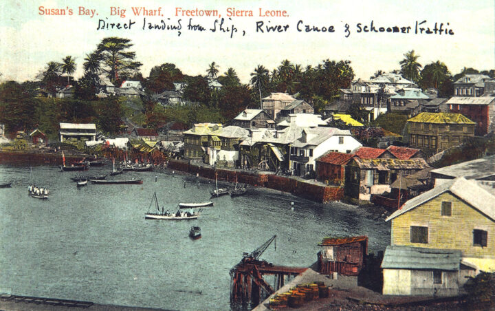 Photograph of houses by a small port. Several boats are cruising in the water. Text printed on the photograph reads "Susan's Bay. Big Wharf. Freetown, Sierra Leone." Handwritten text underneath says "Direct landing from ship, river canoe & schooner traffic".