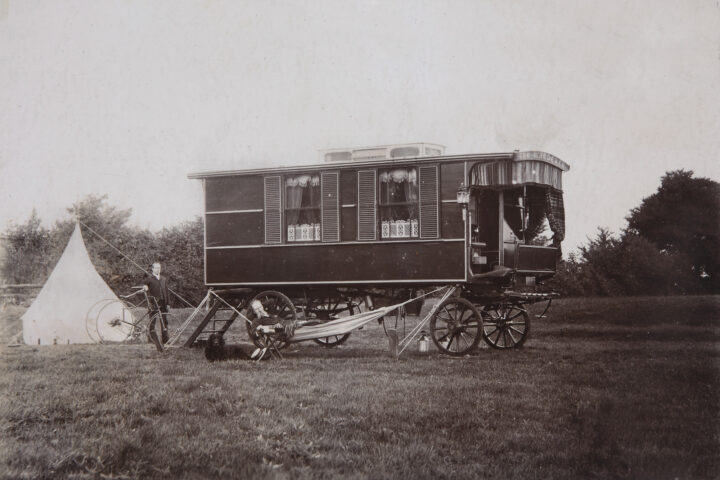 Black and white photograph of an old-fashioned caravan. A man lies in a hammock strung up outside the caravan and another man stands behind the caravan holding a bike.