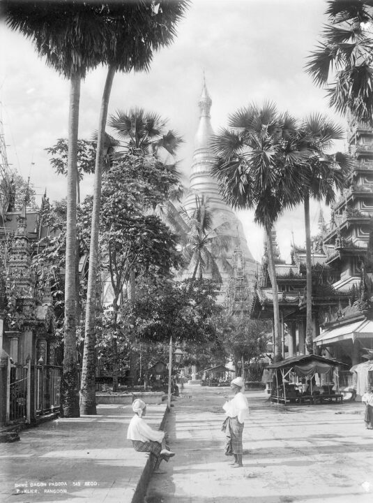 Black and white photograph of a street lined by palm trees with a large pagoda in the background. Two people talk in the foreground, one standing and one sitting on the sidewalk.