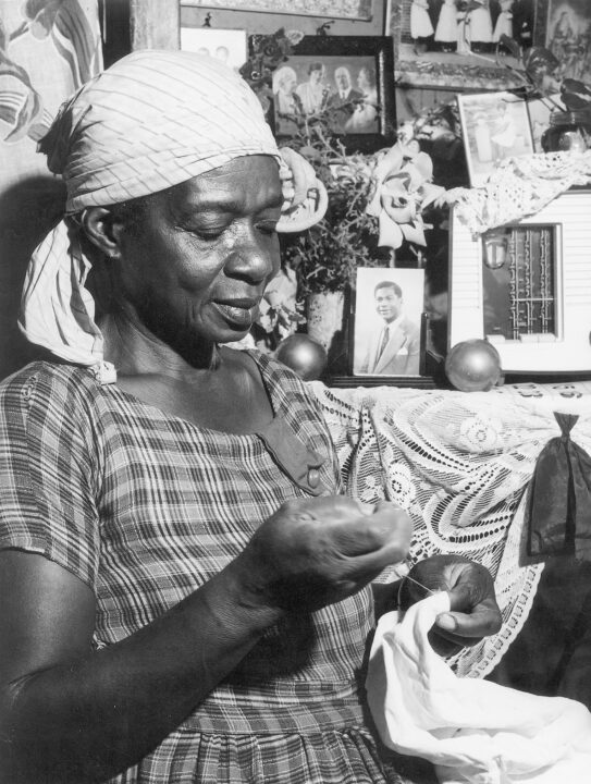 Black and white photograph of an Afro-Caribbean woman sewing as she listens to a radio on the table next to her. She is surrounded by family photos and decorative objects.