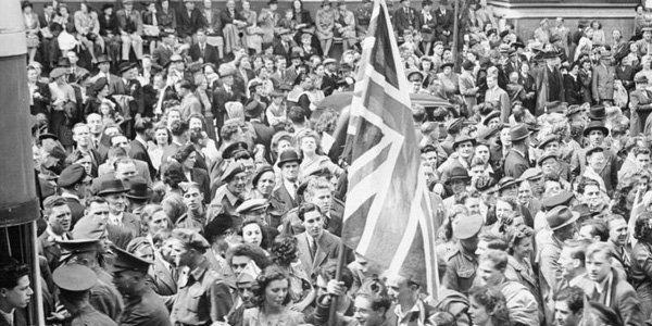 A young man holds aloft a large Union Flag amongst the huge crowd of people that has gathered in Whitehall to listen to Churchill's Victory speech and to celebrate Victory in Europe Day. The crowd is a mix of service personnel, civilians and children. To the left of the photograph, the back of a bus, also packed with people, is just visible.