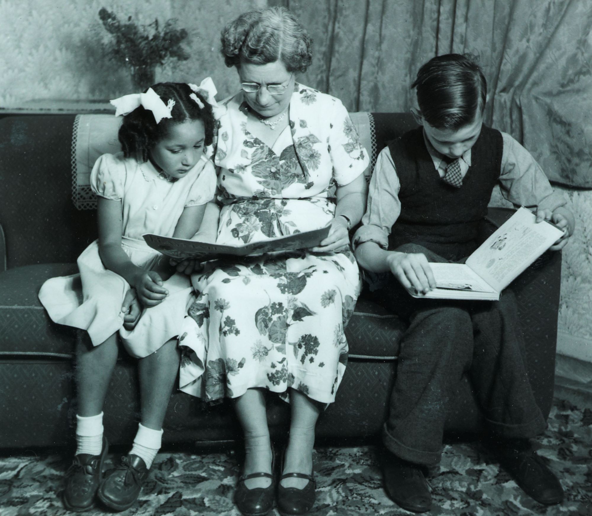 A 1950s foster mother sitting with a girl and a boy on a sofa, teaching them how to read