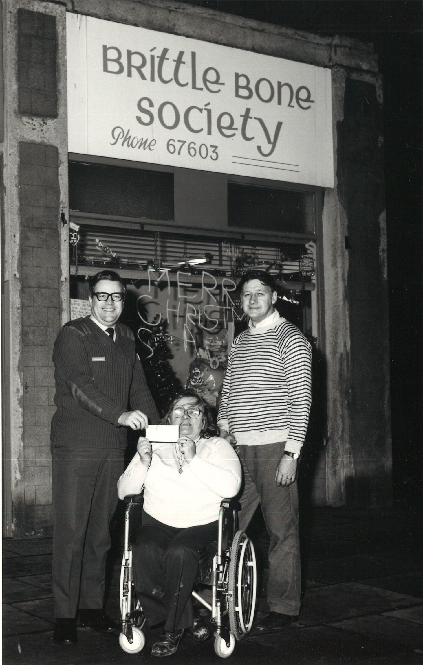 A person in a wheelchair in front of a Brittle Bone Society shop, with a person standing on either side of them