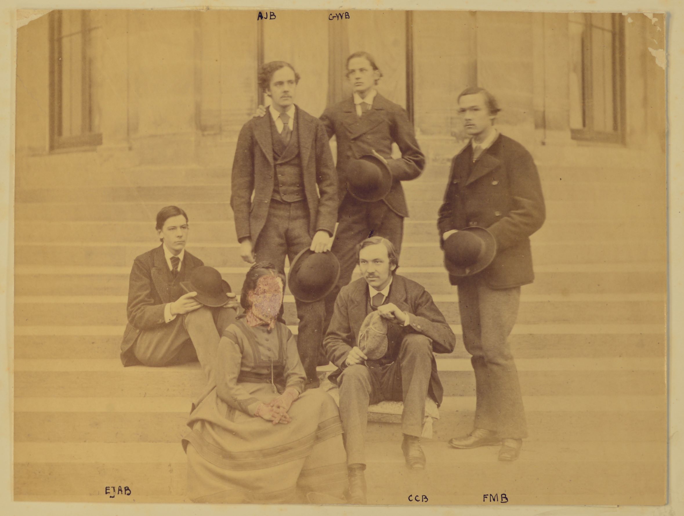 A sepia photograph of four men and a woman, whose face is scratched out, on some outside steps