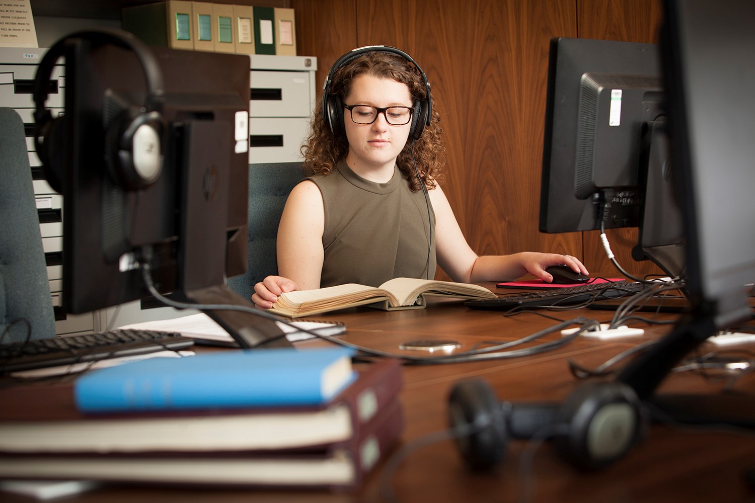 A woman at a desk with headphones on sat at a desk with a computer and book