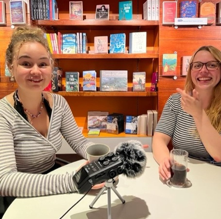 Two women sat a table with a microphone in front of a wall of books