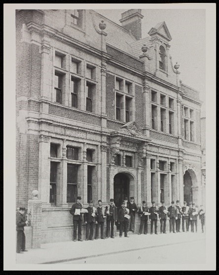 A faded black and white image of a group of men in uniform, standing outside a large brick and stone building.