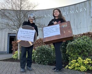 A pair of students stand outside the wooden panelled Inmans Primary School. The one on the left is wearing a helmet and holding a shield, with a white piece of paper attached to it which reads: 'We're really excited to see where our Local History Project takes us!' The student on the right is holding a suitcase, also with a piece of white paper attached, which reads: 'Thank you National Archives for supporting our Inmans Local History Project.'