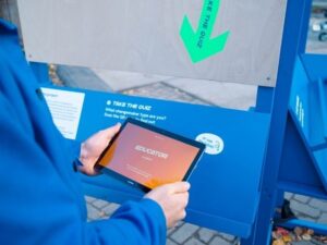 Looking over the shoulder of a person in a blue jumper, holding an electronic tablet that is displaying the word 'Educator' on screen. In front of them is a stand, with a downward arrow that reads' take the quiz', pointing to a QR code and the words 'or use the tablet'.