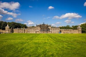 A grand, stone-built stately home, viewed from across a large grass lawn. Blue sky is above, dotted with small clouds.