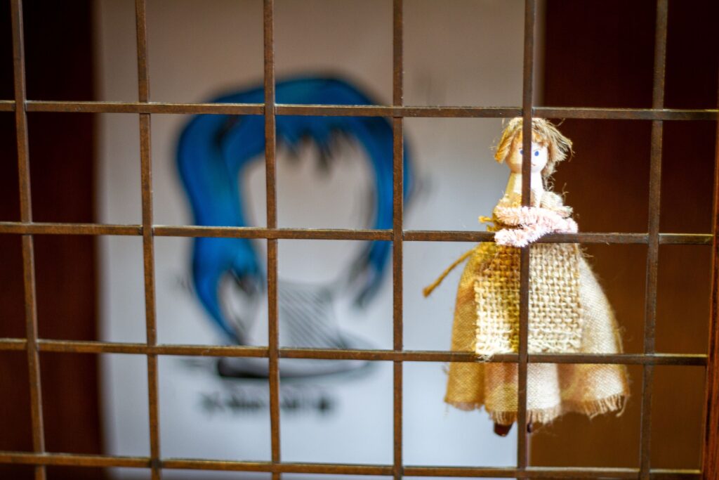 A copper-colour grating, which has a small handcrafted doll attached with pipe cleaners, which double as the doll's arms. An illustration of a blank bust is in the background, with shoulder-length bright blue hair.