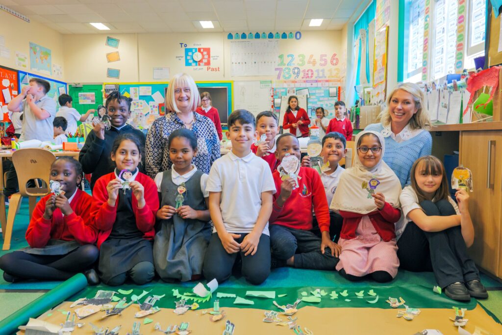 Interior of a Primary School classroom - in the foreground, a group of 10 children, wearing red, white and grey school colours, are kneeling or sitting on the floor as they pose for a photo, each holding a paper figure. Two adults are sat on the floor behind them.