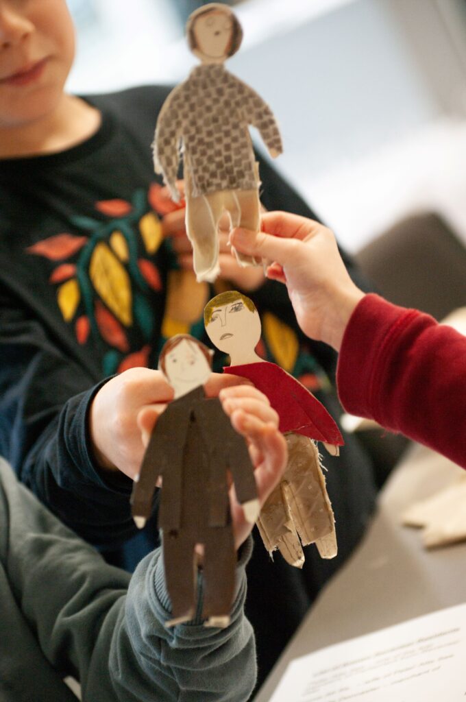 A close-up of three hand-created paper and fabric figures, each being held by a young child