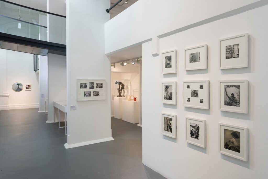 A white gallery space with a grey floor, with nine white-framed black and white photographs in the foreground, and a few sculptures in the background.