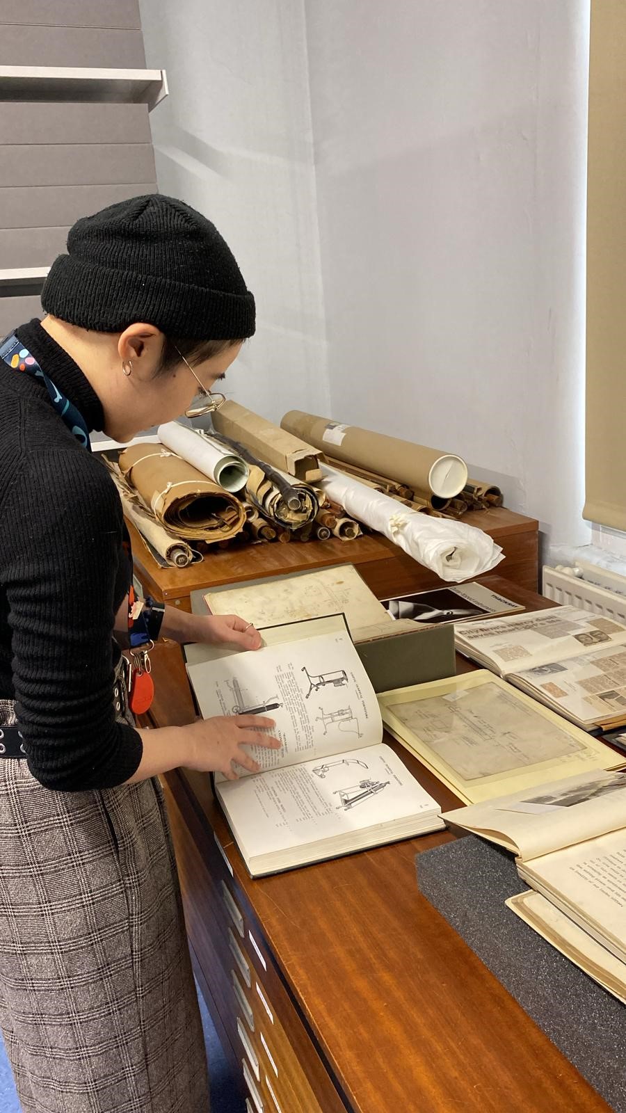 A person standing at a large wooden plan chest, handling a small hardback book with diagrams in it. Around them are various books, and on a cabinet to the left of them are several rolled documents of various sizes.
