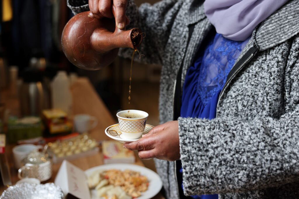 A person wearing a blue top, a lilac scarf and a grey fleece jacket pours a golden brown liquid from an ornate terracotta-colour pot into a small white and gold china cup. They are resting the cup on a matching saucer, held in their other hand.
