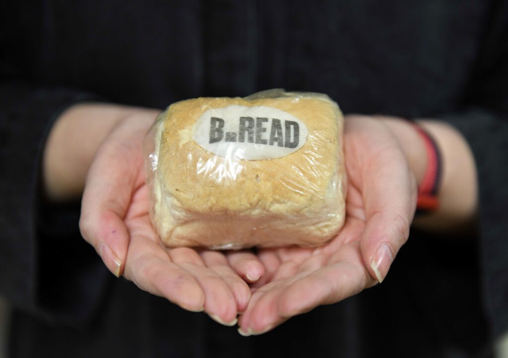 Close-up of a very small loaf of bread, wrapped in clear film. A sticker on the bread reads 'B.READ'. The loaf is held in the cupped palms of a person, who is wearing black.