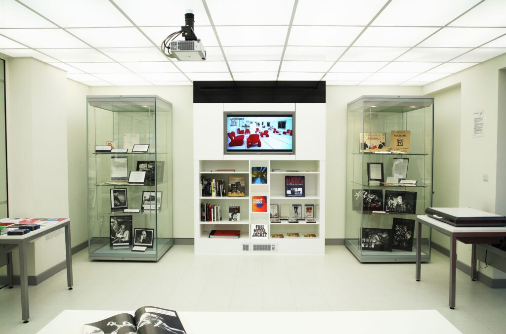 A white square room, where the focus is the far wall. There are two tall glass display cabinets either side of a central, low, white in-built bookcase. Above the bookcase is an inbuilt TV monitor, that is displaying a picture of some red chairs in a white room