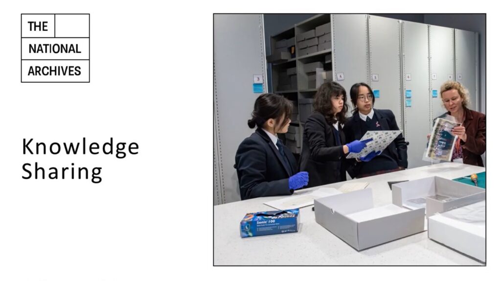 White presentation slide, with 'Knowledge Sharing' typed in black on the left of the page, halfway down. The National Archives logo is top left. On the right, just over half of the slide is taken up with an image of three students in dark blue blazers, wearing purple disposable gloves, and standing in an archive storage space with high shelving. The middle student is holding a document