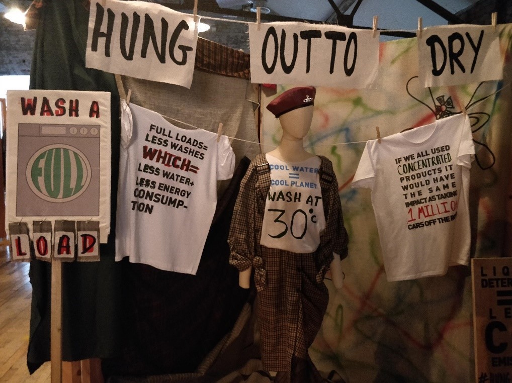 Mock washing line, with 'Hung out to dry' written in thick black marker on beige cloth pegged to the line. A lower line is in front, with two white t-shirts with slogans printed on them. The first reads 'Full loads = less washes which = less water + less energy consumption'. The second t-shirt slogan reads 'If we all used concentrated products it would have the same impact as taking 1 million cars off the road'. In between the t-shirts is a mannequin wearing a brown check dress and a burgundy beret. The mannequin also has a white bib with a handwritten slogan, which reads 'Cool water = cool planet: wash at 30 degrees'. To the left of the t-shirts and the mannequin is a mock protest sign that reads 'Wash a full load'.