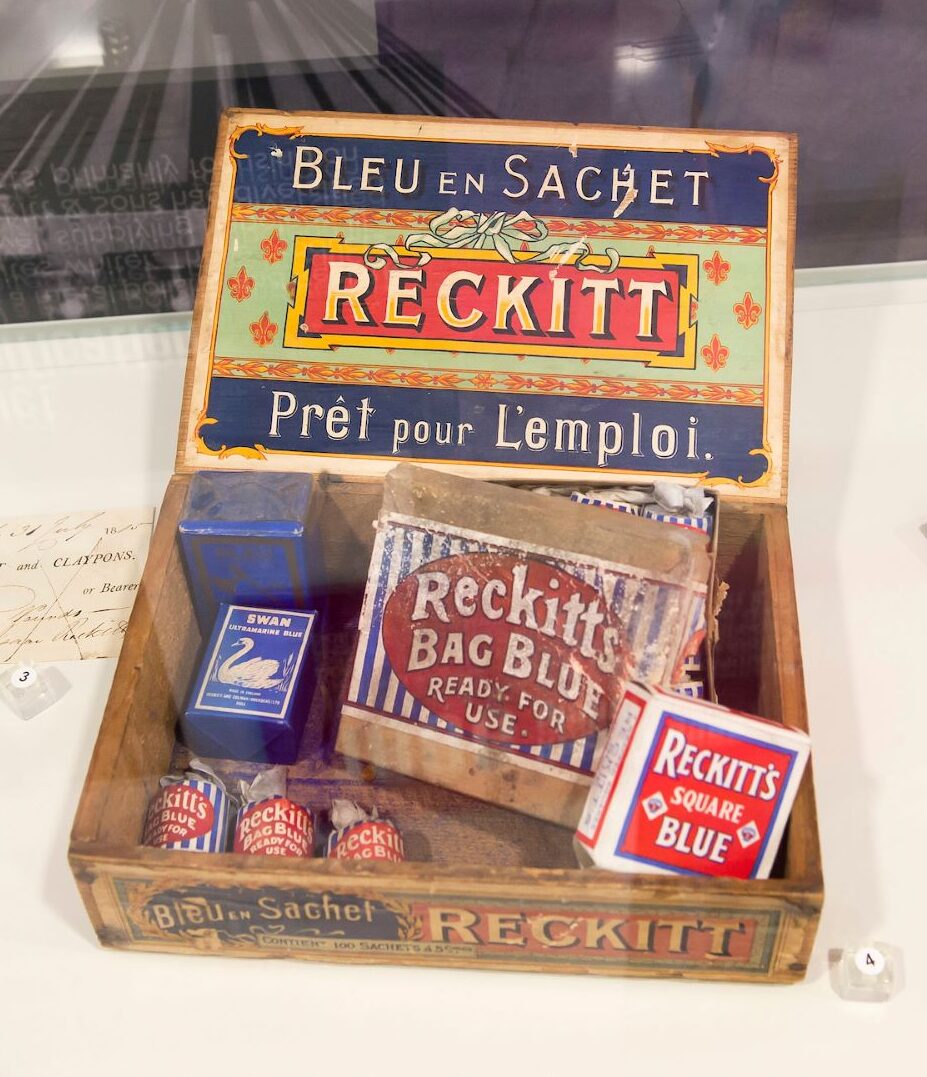 An open vintage wooden box, with a colourful printed design on the inside of the lid that reads 'Bleu en Sachet - Reckitt - Pret pour L'emploi.' In the box a vintage items - two small packs of Swan Contemporary Blue, a large tin of Reckitt's Bag Blue, a smaller box of Reckitt's Square Blue, and three small tubes of Reckitt's Bag Blue.