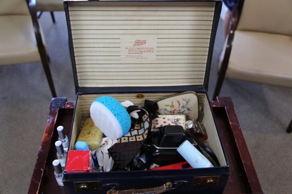 An open black suitcase, on a dark wood table, with items inside, including: a two-sided baby blue and white sponge, two patterned ties, a camera, a pair of cream-coloured gloves, and a small perfume bottle