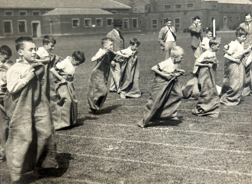A black and white photograph of around a dozen boys taking part in a sack race on a school field.