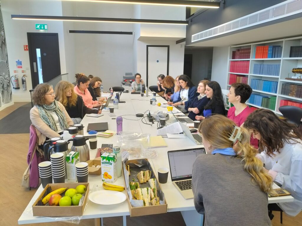 A dozen people sit around a long table having a discussion. Many of the people have laptops open. There are snacks and paper cups on the table also.