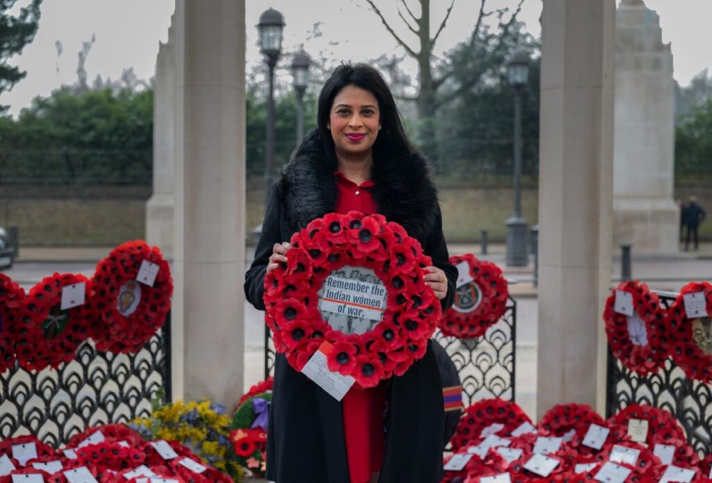A woman stands between two concrete pillars holding a wreath of poppies. She is surrounded by more wreaths with cards attached.