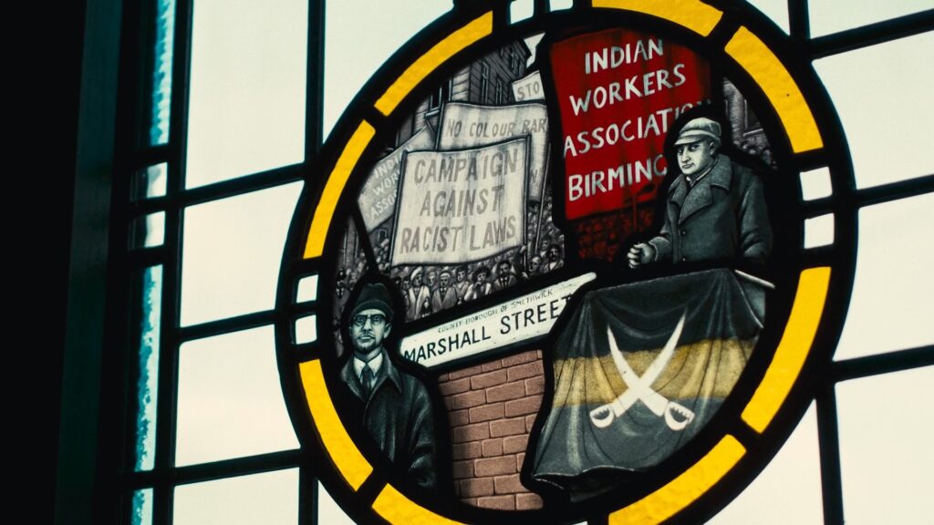 A stained glass window depicting a crowd of people marching through a street behind a brick wall with the sign 'Marshall Street'. The crowd holds up banners reading 'Campaign Against Racist Laws' and 'Indian Workers'.