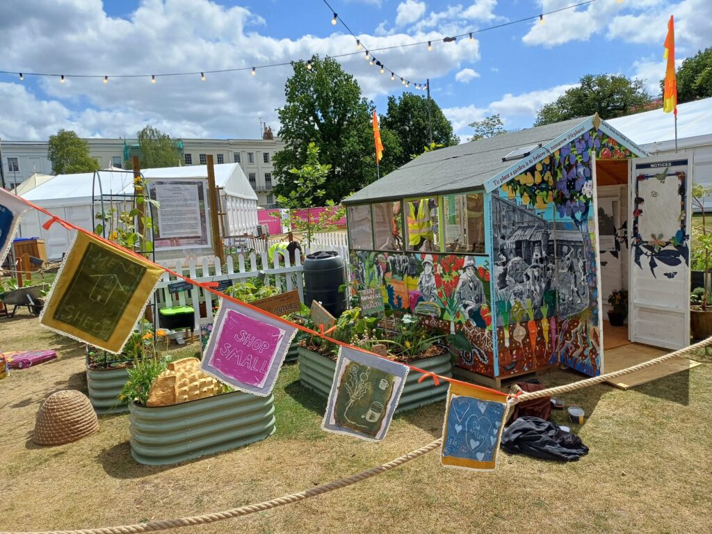 An open shed painted entirely with colourful illustrations, surrounded by gardening beds and a display of artworks hung on suspended rope.
