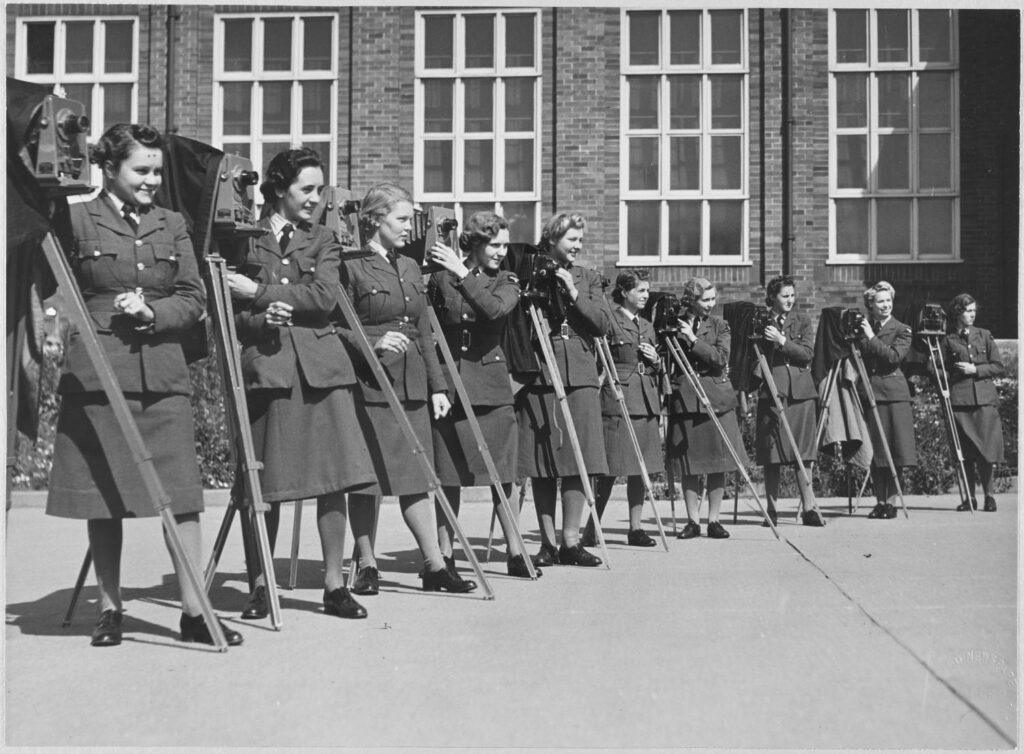 A black and white photograph showing a group of women in military uniform standing in a line. Each one stands next to their own tripod-mounted camera.