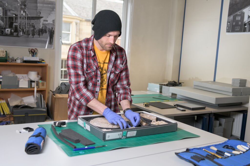 A man in a conservation studio carefully handles a box of items, wearing latex gloves.