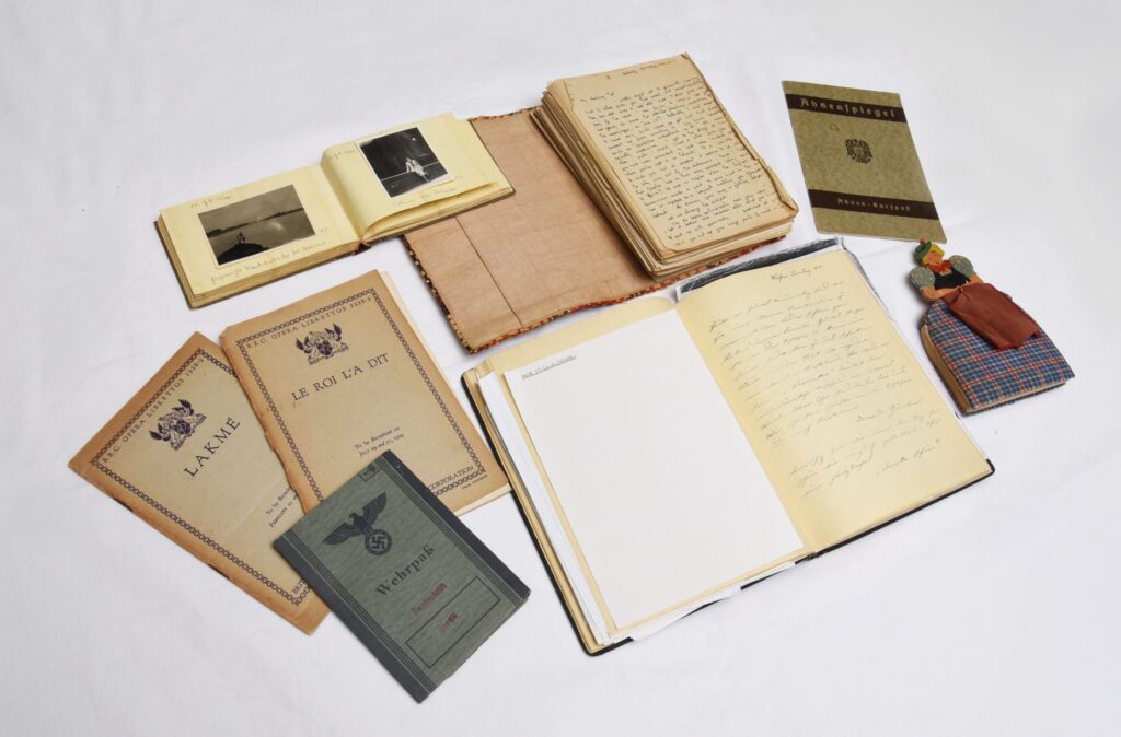 A selection of toned documents and items laid out on a white surface. Most of these appear to be diaries or identification documents. There is also a thread stitching of a woman in a long dress.