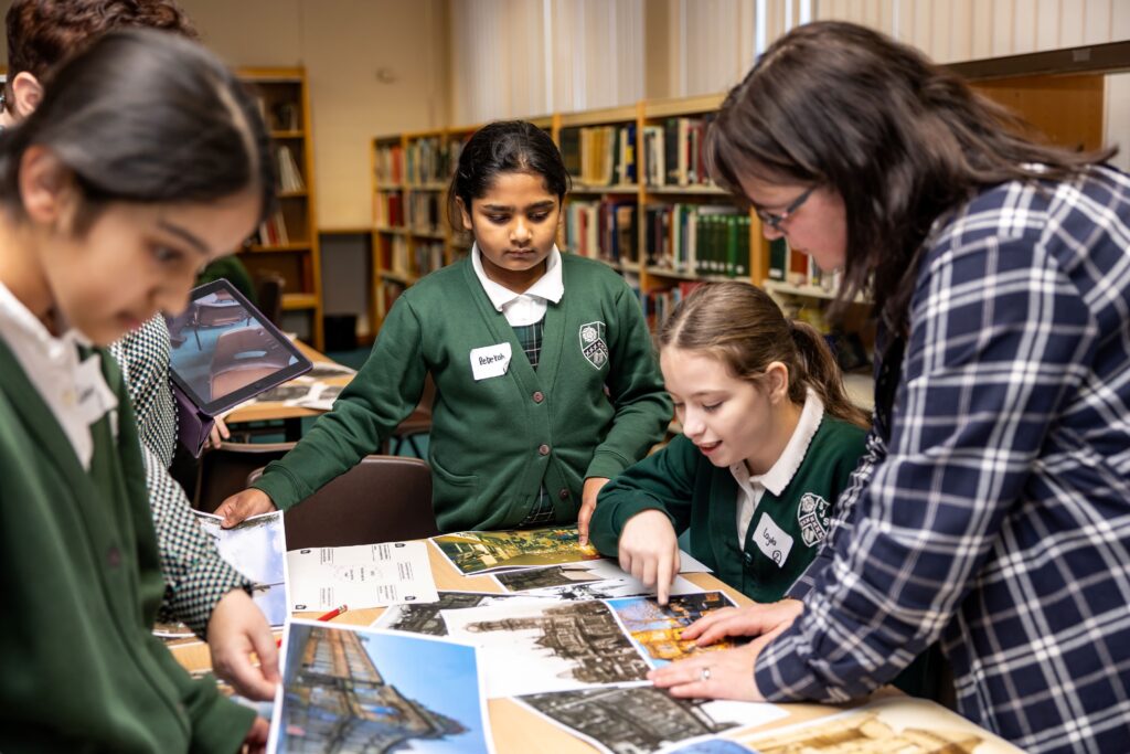 A small group of school children, and an adult, examine a series of photographs depicting buildings. Book shelves are lined up behind them. 