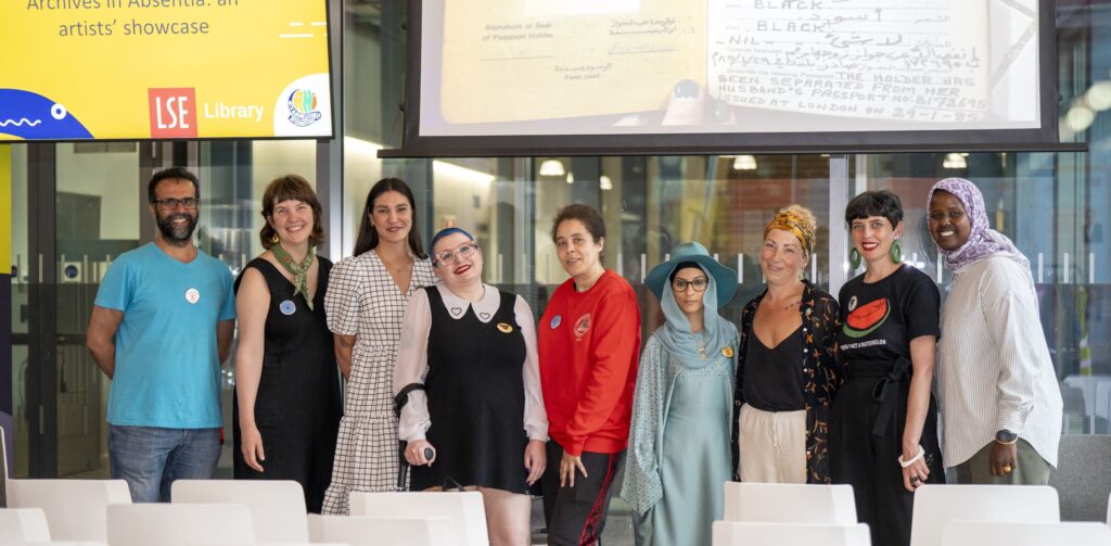 A group of nine people stand in a row in a large room, with rows of chairs in front of them and a screen and projector behind them.