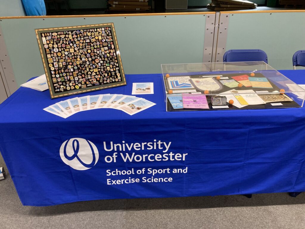 An exhibition table stand holding flyers, documents displayed in a glass case and a framed display of badges. The table cloth reads: 'University of Worcester'.