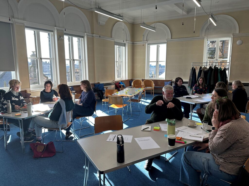 Around a dozen people sit on tables in groups of three or four having discussions with papers and pen laid out on each table.
