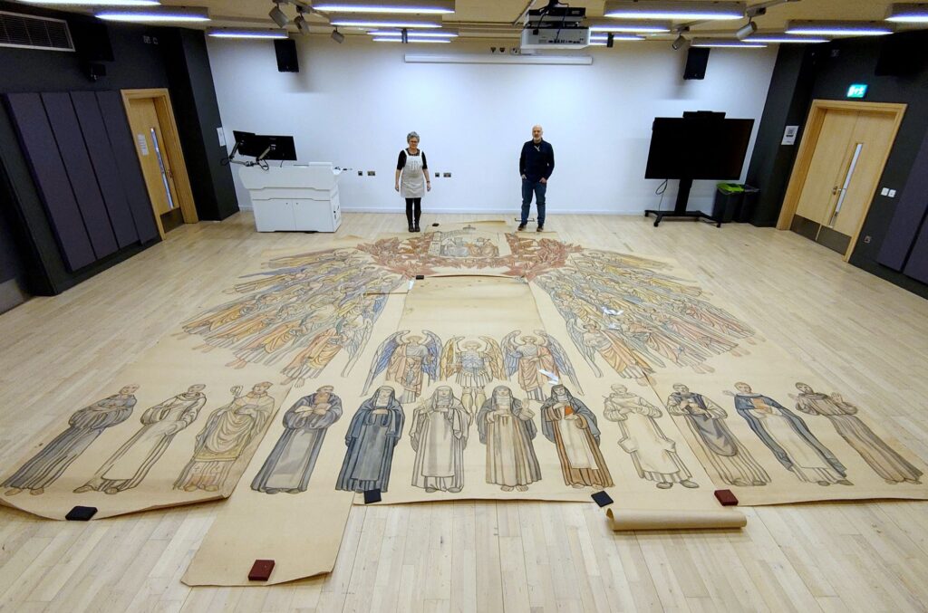 Two people stand in a large empty room looking down at a giant paper document showing a choir of angels that is laid out on the floor.