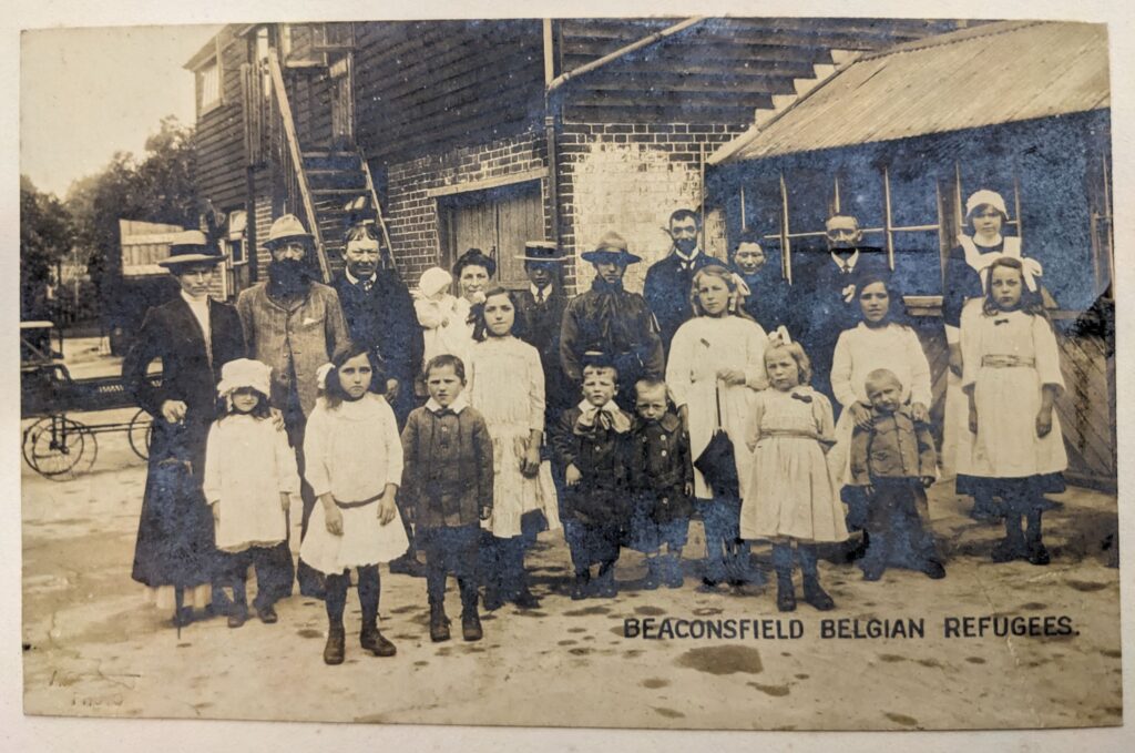 A sepia photograph entitled 'Beaconsfield Belgian Refugees' of a group of men, women and children standing in a yard in front of a brick and wooden building