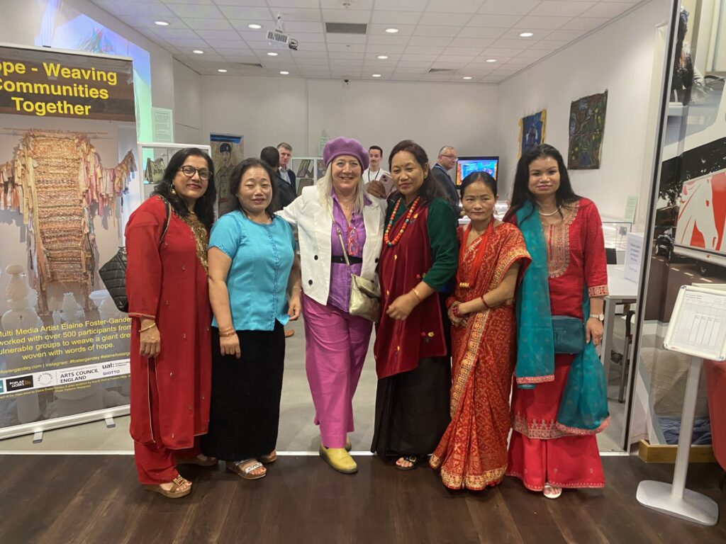 Several woman stand in a line posing for the camera. Behind them we see an exhibition space and a banner that reads 'Weaving communities together'
