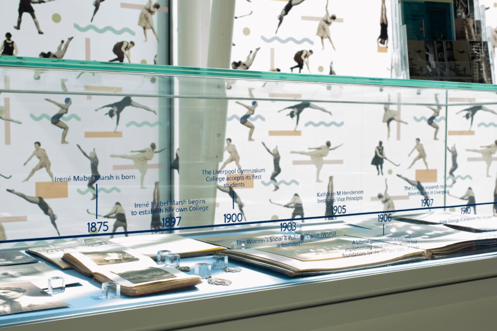 A glass exhibition display of books and documents. Printed on the glass are icons of people playing sports, along with dates and small smatterings of text.
